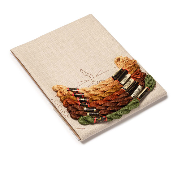  DIY embroidery kit for a pinecone table scarf design, showing folded natural linen stamped with pinecones alongside arranged embroidery thread skeins in a variety of colors.