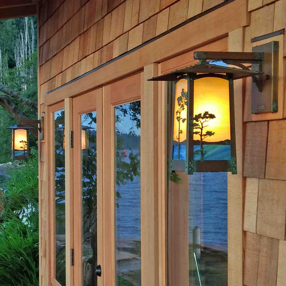  A pair of rustic, artistic arm mounted wall sconce light fixtures with peaked roof, Monterey Pine artwork, an aged patina finish and golden glass installed on a shingle home next to a lake in the woods.