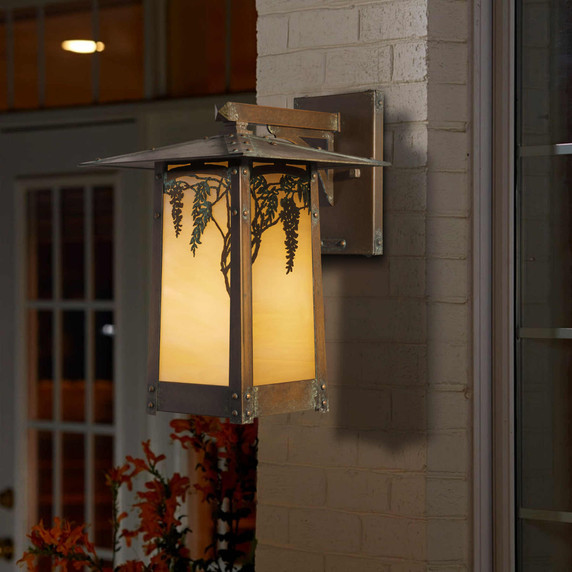  A rustic, artistic arm mounted wall sconce light fixture with wisteria artwork, an aged patina finish and golden glass installed on a painted brick wall near windows.