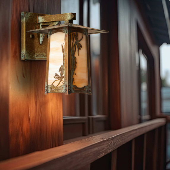  A rustic, artistic arm mounted wall sconce light fixture with a peaked roof, dragonfly artwork, an aged patina finish and warm white glass installed on an exterior wall of a home.