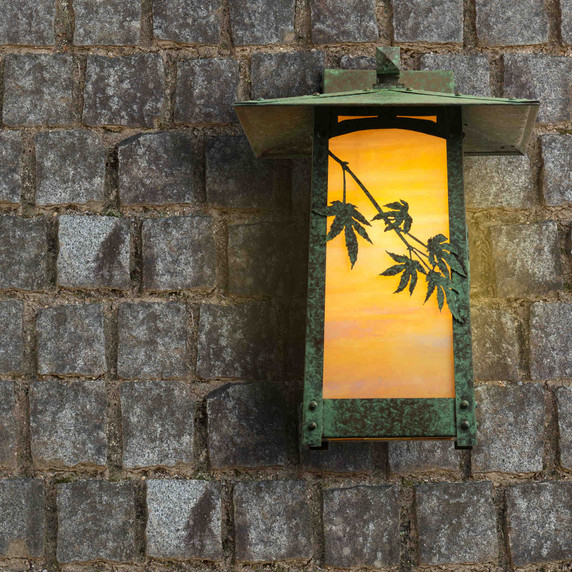  A rustic, artistic flush mounted wall lantern with a peaked roof, Japanese Maple artwork, a verdigris finish and iridescent glass installed on a stone tile wall.