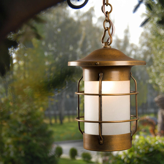  A nautical, streamlined art deco chain pendant lamp or lantern with an aged brass finish and bubbly white glass hanging from a backyard porch with a view of the landscaping.
