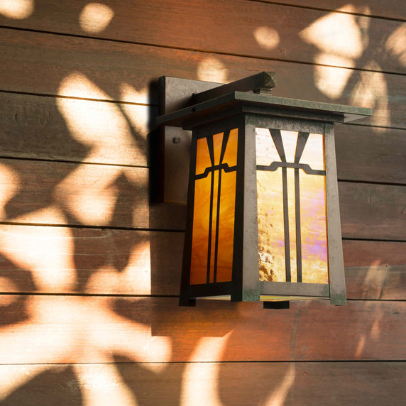  An Arts & Crafts wall mounted sconce light fixture with an aged bronze finish and iridescent glass installed a a wood plank wall with heavy light and shadow contrast.