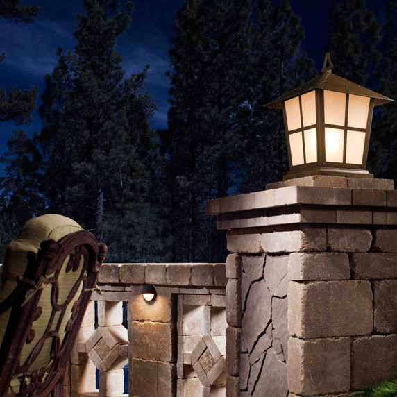  A classic, traditional sloped-roof column mounted pier light fixture with crown molding accent, an aged bronze finish and warm white glass installed in a luxurious backyard on a stone wall.
