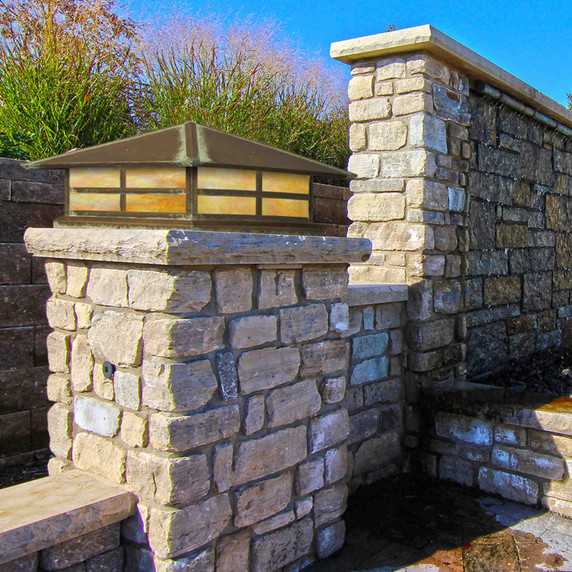  A Prairie Style column mounted pier light fixture with a window pattern, aged patina finish and iridescent glass installed on a stone pillar near a flower box in the backyard of a large stone house.