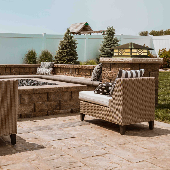  A Prairie Style column mounted pier light fixture with a chevron pattern, an aged patina finish and warm white glass installed on a stone pillar next to a firepit and patio furniture in a backyard.