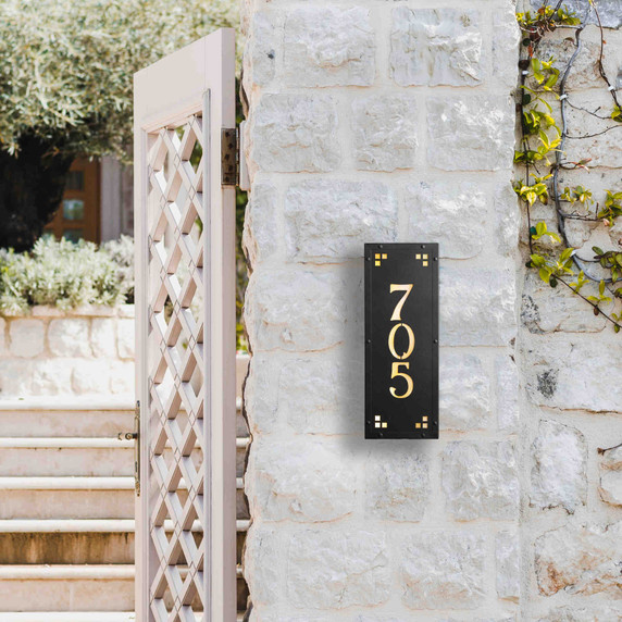  A solid brass, vertically oriented illuminated house number plaque with a three number address, a satin black finish, warm-toned glass and corner glass accents. It's installed on a white stone wall next to a white metal gate.
