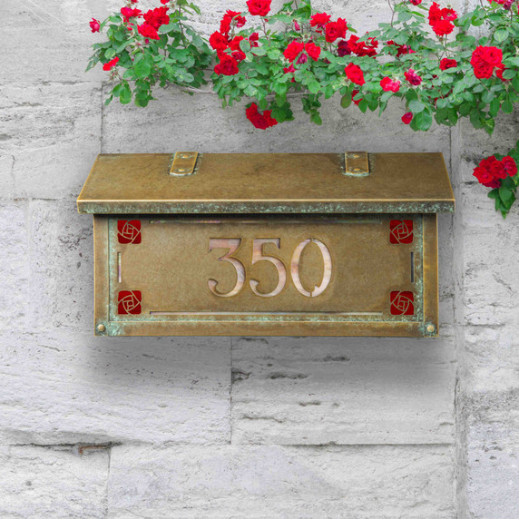  A Craftsman style mailbox featuring the Hunter Rose (sometimes called the Mackintosh Rose) as corner design accents. The mailbox has an aged patina finish and house address numbers backed by iridescent glass. It's installed on a stone wall under climbing red flowers.