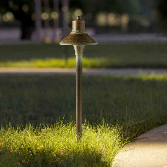  A garden or pathway landscape light with a flared top, aged patina finish and a decorative round finial topper lighting a winding section of path at dusk.
