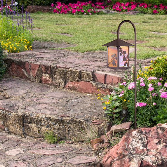  An artistic garden lantern on a shepherd's hook post with a smooth bronze finish, ivy artwork and iridescent glass installed in a flower bed next to stone steps.