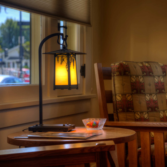  A rustic, artistic table lamp on a hooked arm. The lantern has a peaked roof, wisteria artwork, an oil-rubbed bronze finish and golden glass. It sits on a circular side table with a candy dish next to a Craftsman style Stickley easy chair.