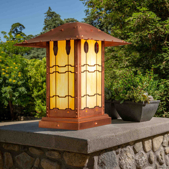  An authentic reproduction of backyard patio column landscaping lantern at the Gamble House in Pasadena, California.The pier mounted fixture is illuminated and has a raw copper finish and custom glasswork. It is installed on a stone platform with potted plants nearby and flowering shrubs in the background.