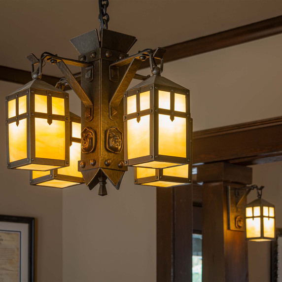  A unique, Arts & Crafts style chandelier with four lanterns, a medieval aesthetic and a cast friar's head facing each direction on the body of the fixture. It has an oil-rubbed bronze finish and golden glass. There is a matching sconce visible in the background.