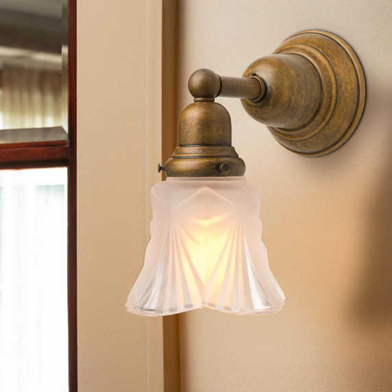 A reproduction Craftsman style bungalow wall sconce light fixture installed next to a beveled glass window. The sconce has an aged brass finish and an Art Deco glass shade with a drapery pattern. The shade is aimed downward.