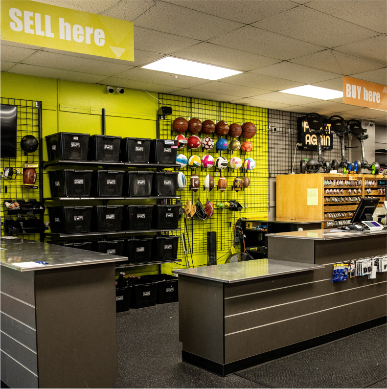 The front counter of a sporting goods store with a shelf behind the counter full of sporting gear like soccer balls, basketballs, and baseball gloves.