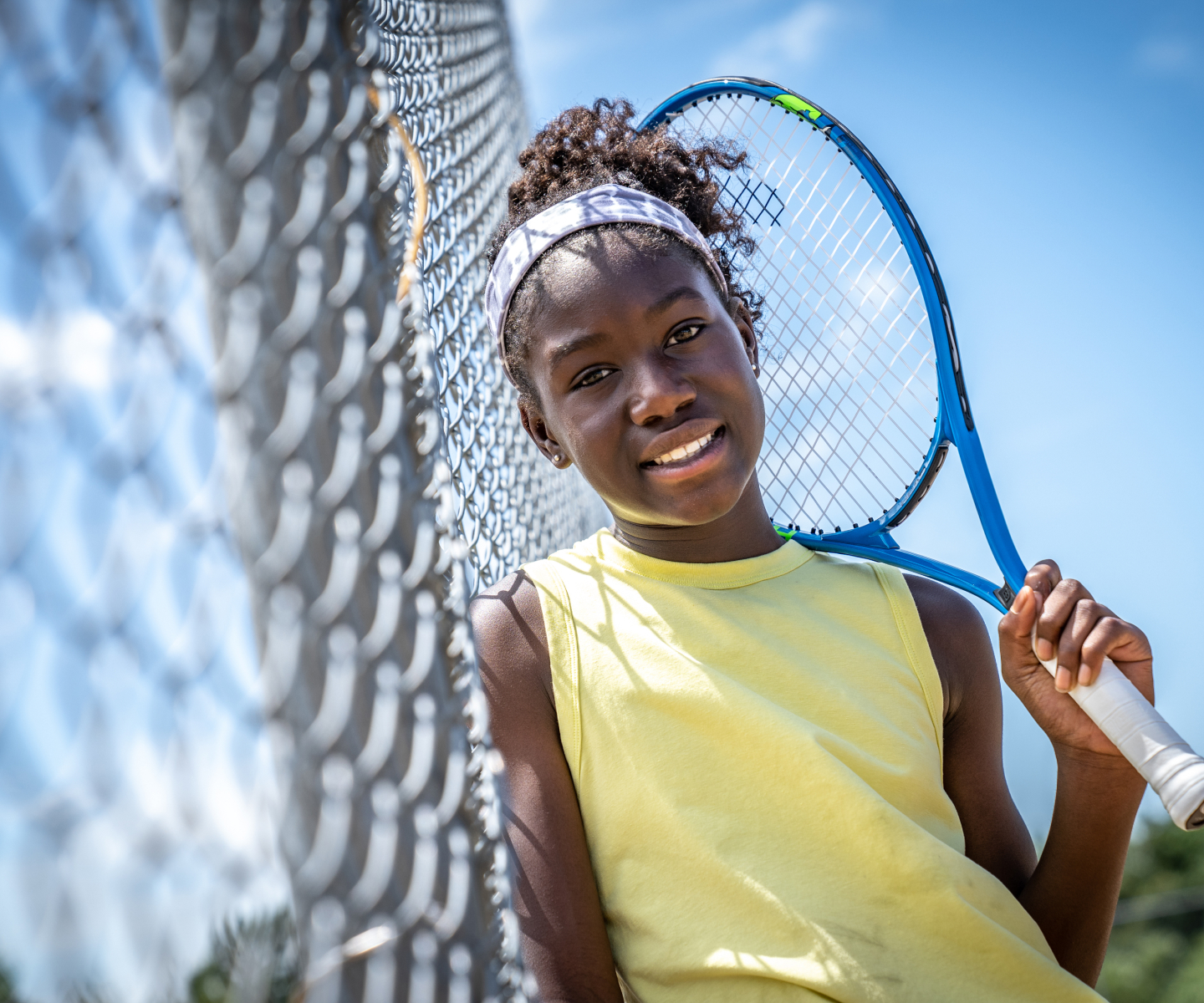 young female tennis player leaning against a chain link fence holding her racket and smiling