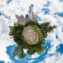 A 360-degree panoramic Spherescape of Central Park South taken from a bedrock outcropping, showcasing the natural rock foreground with a backdrop of skyscrapers and trees against a cloudy sky.