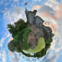 A 360-degree panoramic image of Belvedere Castle in Central Park, NYC, taken at sunset with vibrant sky colors and surrounding greenery, reflected in the waters below in a spherical composition.