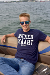 Model wearing navy Wicked Smaaht Boston t-shirt with white collegiate lettering while sitting on a boat in Boston Harbor.