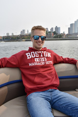 Man wearing a cardinal red Boston 1630 Massachusetts hoodie with white distressed screen-printed lettering, sitting on a boat with Boston skyline in the background.