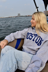 Woman wearing a sport grey Boston hoodie with navy collegiate lettering while sitting on a boat with Boston waterfront skyline in the background.