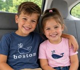 Youth Boston T-shirt featuring a playful spouting whale graphic and "boston" lettering, available in Heather Navy and Light Pink.