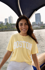Butter yellow Comfort Colors Boston T-shirt with white collegiate lettering displayed on model by the Boston waterfront.
