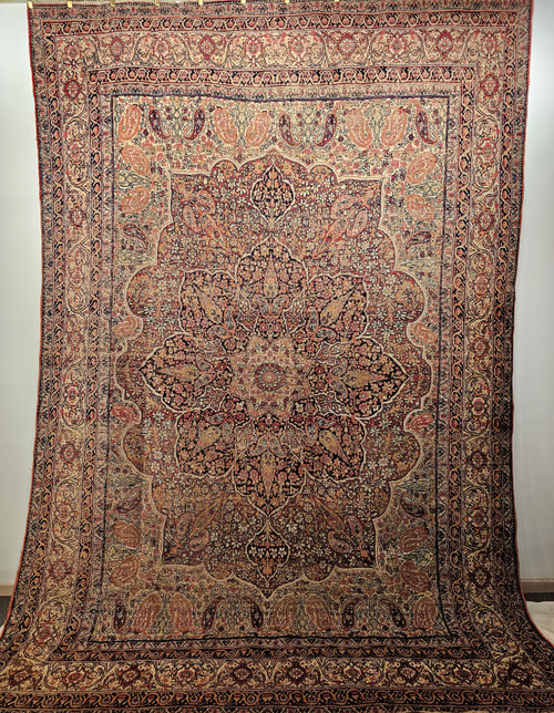 Late 19th Century Antique Persian Kerman Lavar room size rug. Hand-woven. Floral and paisley design. Ivory background. Central medallion in navy blue and caramel. Paisley forms in red and pink. Cypress trees in pale green. Colors: ivory, red, pale yellow, midnight blue.