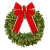 This image shows a festive green Christmas wreath decorated with warm white lights and topped with a large red bow featuring gold trim. The bow's long ribbon tails cascade down the front, adding a classic holiday touch to the arrangement