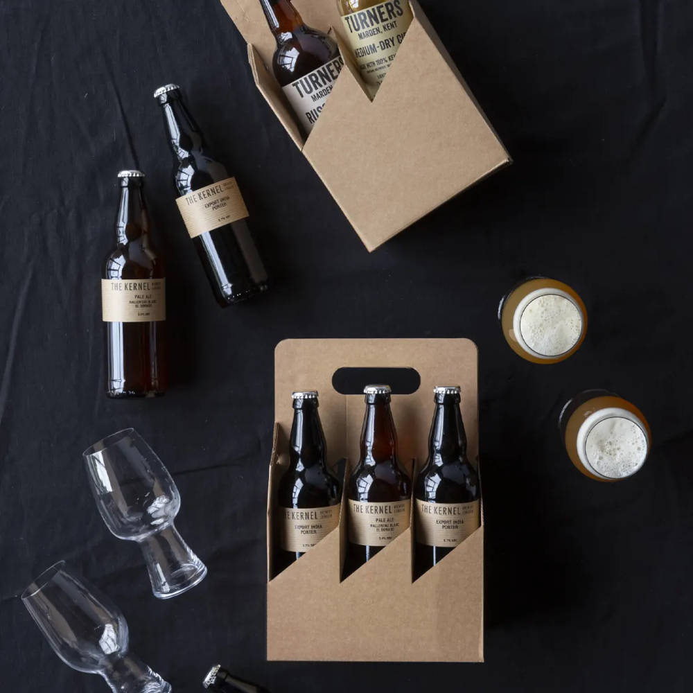 A box containing three beer bottles on a black background with additional bottles and glasses around it