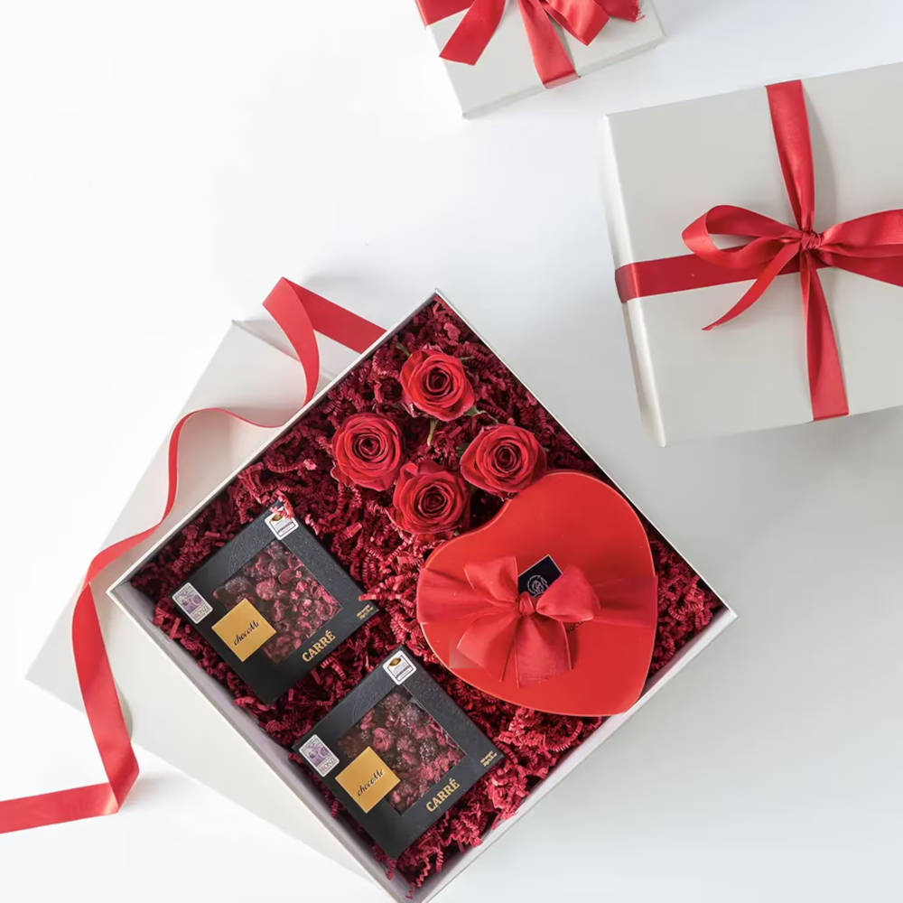 An open box with a heart shaped box inside, two other small boxes and four rose heads on a bed of red paper