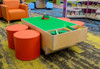 Playscapes 3-Branch Discovery Lego Table, indoor children’s play area with green LEGO building surface, surrounded by orange seating stools and colorful furniture.