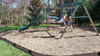 Frame It All Weathered Wood Straight Playground Border Kit 64' - 1" Profile, outdoor playground with children playing on swings and slide, surrounded by trees and grass