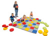 Novum Let's Play Colors Floor Game, children playing on a colorful mat with large circular game pieces in red, yellow, and green, overseen by an adult in a gray hoodie.