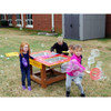 Infinity Playgrounds Bubble Station Activity Table, children playing with bubbles outdoors in a grassy area with building and storage shed in the background.