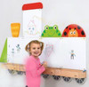 Moje Bambino Toddler Train Magnetic Board Set, a young girl drawing on a magnetic whiteboard with colorful magnetic character pieces above the train panel.