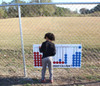 Infinity Playgrounds Connect 4 Playground Game, a child playing with a large outdoor Connect 4 game on a fence outside on a grassy field.