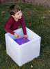 TickiT Sensory Mood Light Play Cube, a white LED light-up cube filled with purple sand and small colorful toys for sensory play, used outdoors by a young boy.