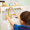 Teamson Kids Versailles Play Kitchen with Refrigerator and Cookware Accessories, a child playing with a miniature kitchen set featuring a golden faucet and sink.