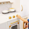 Teamson Kids Versailles Play Kitchen with Refrigerator and Cookware Accessories, a children's play kitchen made of white and gold materials with stove, oven, and oven door, featuring a child's hand adjusting the stove dial.