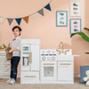 Teamson Kids Little Chef Charlotte Modern White Play Kitchen, a child's play kitchen set with a pretend refrigerator, oven, sink, and stove, featuring gold accents and a digital display, situated in a playroom with framed vegetable illustrations on the wall.