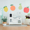 Teamson Kids Little Chef Westchester Ivory Retro Play Kitchen, white miniature play kitchen with stove, oven, microwave, sink, and storage compartments, set up in a child's playroom with colorful fruit wall decals and toys nearby.