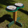 Playmore Designs DouBBle Play Drum Table, outdoor double drum table playground equipment for children, with two drum surfaces on a green and wooden stand on grass Playmore Designs DouBBle Play Drum Table, outdoor double drum table playground equipment for children, with two drum surfaces on a green and wooden stand on grass