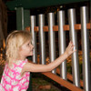 Playmore Designs Outdoor 6-Note Eco Chime Unit, a young girl playing with a metal outdoor chime made of six vertical tubes in a playground setting.