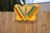 Playmore Designs Butterfly Music Chimes, colorful butterfly-shaped outdoor musical instrument with plastic chimes in red, yellow, blue, purple, green, and orange, mounted on a wooden fence.