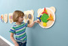 Moje Bambino Spinning Pinwheels Sensory Wall Activity Toy, a young boy interacting with a colorful spinning eco-wooden clock with rotating pinwheels on a classroom wall.