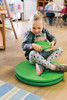The Freckled Frog Whizzy Dizzy Sit 'n Spin, a young girl sitting on a green spinning toy, enjoying playtime in a bright indoor setting.