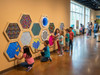 LiteZilla Multi-Sensory Hexagon Panel - Hex-A-Glow XL, a colorful interactive sensory wall with multiple hexagon-shaped panels filled with various tactile and visual elements, in a bright and spacious museum setting with children and families exploring and engaging with the installation.