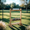 Wood Designs Outdoor Easel for Kids, wooden art easel with a paper roll and compartments full of art supplies outdoors in a grassy park with trees.