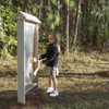 Wood Designs Junior Outdoor Thunderwall, a children’s outdoor educational and play wall with a clear panel and wooden frame, in a forest setting with a young girl engaging with it.